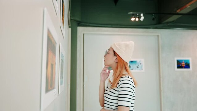 Woman standing she looking art gallery collection in front framed paintings pictures on white wall, Asian people watch at photo frame to leaning against at show exhibition artwork gallery, Side view