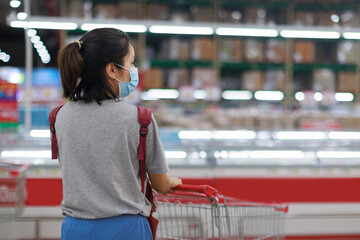 Asian woman wearing face mask and rubber glove push shopping cart in suppermarket departmentstore. Girl choosing, looking grocery things to buy at shelf during coronavirus crisis or covid19 outbreak.