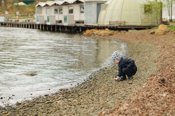 A child boy in a grey hat and dark blue jacket walks near the sea and throws stones at it. Card with selective focus