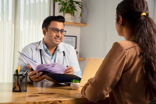 Asian Indian male physician or doctor wearing white apron and stethoscope holding a file of medical report is consulting or prescribing medicines to a Young Female patient in modern clinic or hospital