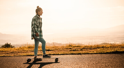 Woman rides at straight road on longboard at sunset time. Skater in casual wear training on board...