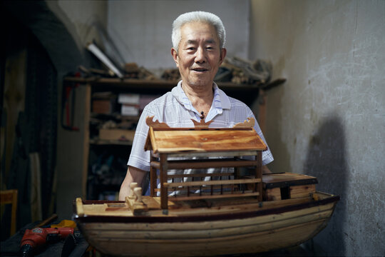 Older Asian Man Works On His Model Boats At His Workshop.