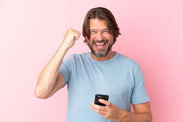 Senior dutch man isolated on pink background using mobile phone and doing victory gesture