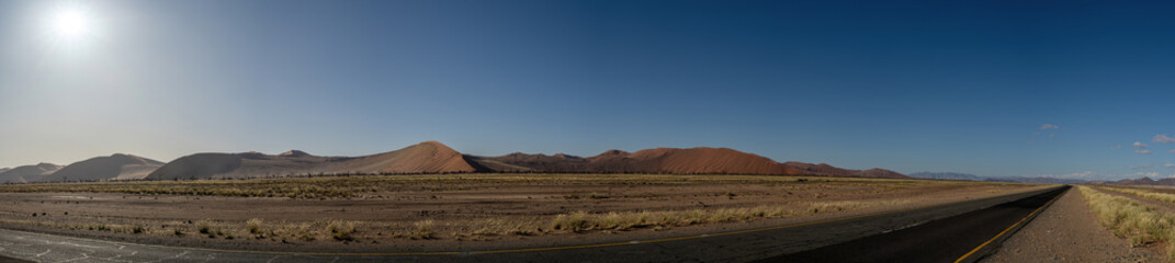 A panorama of dunes in the Namib Desert, on the 
way to Sossusvlei. Road in the foreground with blue sky.