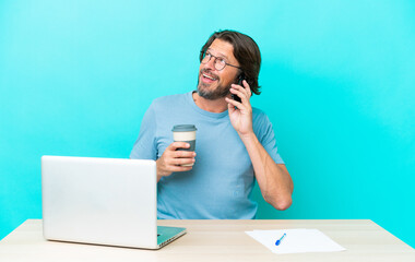 Senior dutch man in a table with a laptop isolated on blue background holding coffee to take away and a mobile