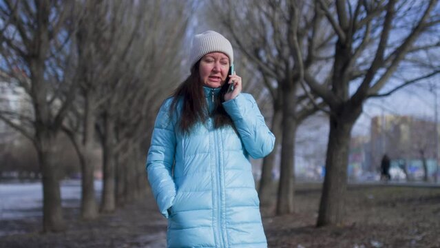 Woman Is Talking On Mobile Phone In Early Spring Park Near The Naked Trees. She Is Wearing Hat And Jacket. Brunette Woman Is Calling On Her Smartphone During Her Walk In City Park. 
