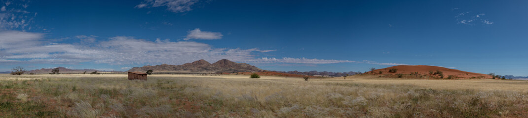 A panoramic photo of hills and mountains in 
central Namibia near Sossusvlei. First red dunes of the Namib Desert visible. Blue sky with scattered blue clouds.