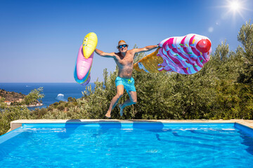 Summer concept with a happy holiday man with hat and sunglasses jumping with two colorful inflatables into a swimming pool