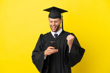 Young university graduate caucasian man isolated on yellow background with phone in victory position
