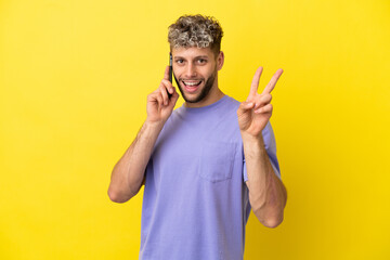 Young caucasian man using mobile phone isolated on yellow background smiling and showing victory sign
