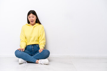 Young Chinese woman sitting on the floor isolated on white wall with surprise facial expression