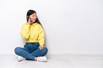 Young Chinese woman sitting on the floor isolated on white wall keeping a conversation with the mobile phone with someone