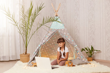 Horizontal shot of charming little girl sitting in wigwam and watching cartoons on laptop, having positive facial expression, looking at notebook screen, enjoying interesting video. © sementsova321