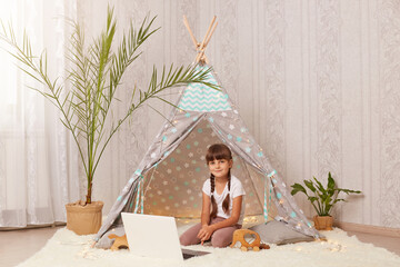 Indoor shot of dark haired little girl sitting in wigwam and using laptop having happy facial expression, looking at camera, enjoying time in peetee tent. © sementsova321