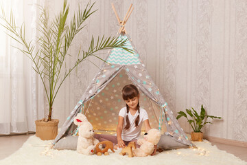 Portrait of Caucasian little girl wearing white t shirt sitting on floor in wigwam and playing with teddy bear and rabbit, talking with her soft toys, enjoying leisure time at home. © sementsova321