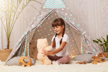 Horizontal shot of smiling satisfied little girl wearing white t shirt sitting on floor in wigwam and playing with teddy bear, holding soft toy in hands, expressing positive emotions. © sementsova321