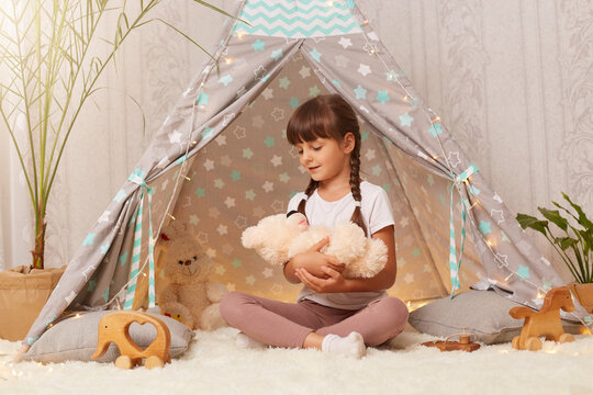 Image of charming little girl wearing white t shirt sitting on floor in wigwam and playing with teddy bear, holding toy in hands, putting her favorite toy to sleep.