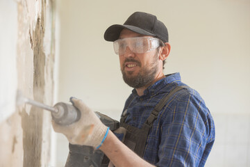 Mature man wearing baseball cap with safety glasses and gloves works on renovation demolition of house uses hammer to tap out tiles in old kitchen bathroom.