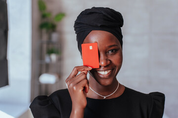 Close up portrait of African cheerful woman holding credit card, covering her face, smiling wide....