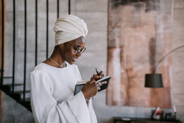 Cheerful African woman in white turban, white dress, glasses holding planner writing notes cute...