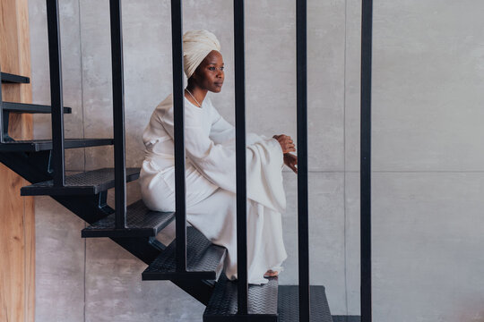 African young adult woman in white turban and white dress sitting on stairs at home looking aside. Calm afro american female relaxing at home in thoughtful mood. Pensive girl thinking about future.