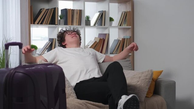 Successful vacation. Satisfied man. Home relax. Happy casual guy in spectacles sitting down sofa with relief near packed suitcase in light room interior.