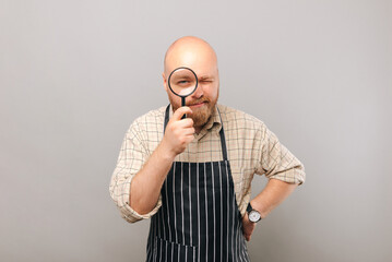 A photo of a bearded young barista holding a magnifying glass being curious near grey wall