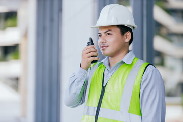 Asian engineer young man wearing safety vest and helmet standing using walkie-talkie for contact work on building construction site background. Engineering construction worker concept.