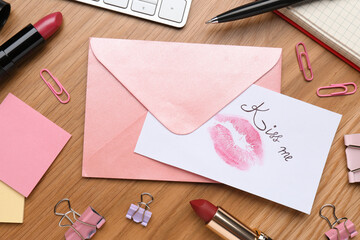 Card with phrase Kiss me, lipstick mark and envelope on wooden office table, flat lay