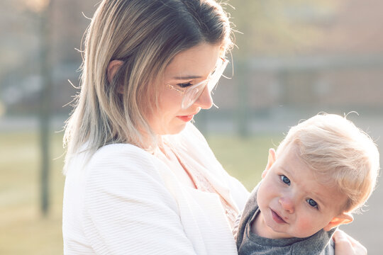 Happy young mother with a small child or toddler in her arms, hugging and cuddling, standing in the middle of a park in the rays of the rising morning sun. High quality photo