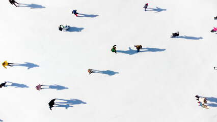 Many people are skating on a white outdoor ice rink in the city on a sunny winter day. Shadows of people skating on the surface of a white ice rink. Aerial drone view. Top view. Lifestyle, sport, rest