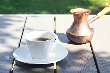 A cup of coffee stands on a wooden table in the garden. Behind is a Turk with freshly brewed coffee.