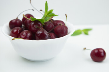 A white plate with a ripe burgundy cherry and a mint petal stands on a white background, there is a cherry and mint leaves next to it, a close-up side view. High quality photo