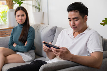 Young Asian couple in which the man plays the game and ignores his wife. His wife's face was filled with indignation. - obrazy, fototapety, plakaty