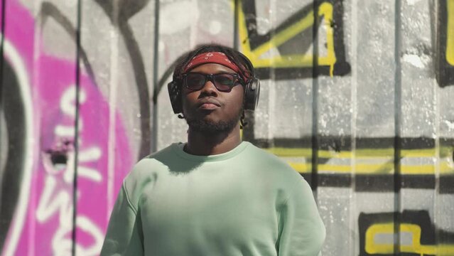 Waist up portrait of young African American hipster man in red bandana and dark sunglasses nodding his head while listening to hip hop music un headphones, standing against graffiti wall outdoors
