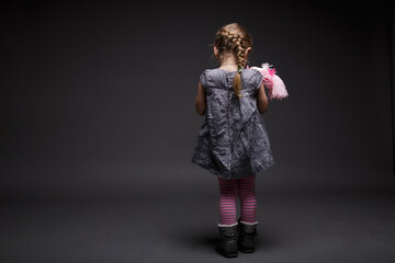 Rear image of a little girl with doll, toddler, offended by someone, being in bad mood, posing isolated over dark grey background.