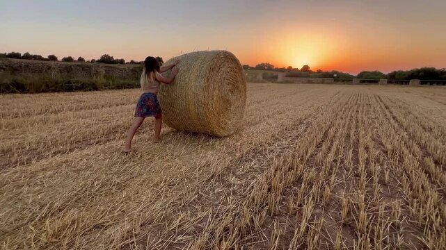 Chica trabajando en el campo haciendo rodar una bala de paja en atardecer de verano