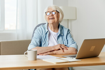 a happy elderly woman with gray hair is sitting at her desk with a laptop and smiling broadly looks away with her hands folded on the table. The concept of working from home