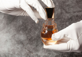 Hands in white gloves holding glass perfume bottle on a gray background