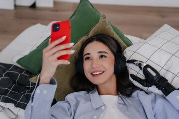 Cheerful young girl with disability laying on bed at home, holding phone by  bionic hand...