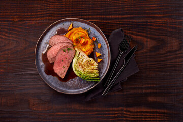 Rinderbraten mit Wirsing und Kürbis auf Holzfond und grauem Teller mit Servieltte und Besteck
