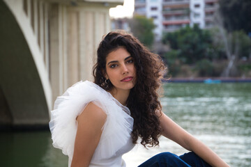 young, beautiful woman with dark, curly hair, wearing a cap, sitting by a river. The woman is posing for photos. Concept expressions. smile, sad, thinking, enjoying, living.