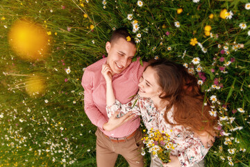 Romantic couple of young people lying down on grass in field of spring flowers. Woman laying on the shoulders of man and looking happily. View from above.