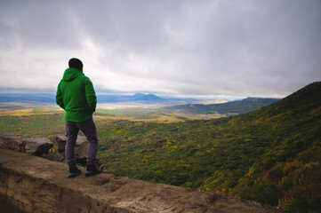 a man traveling on foot, standing in a bright jacket on top of one of the mountains, looking into the distance at the mountains and the cloudy sky