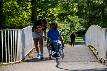 Teenage girl (16-17) in wheelchair with friend walking in park - obrazy, fototapety, plakaty