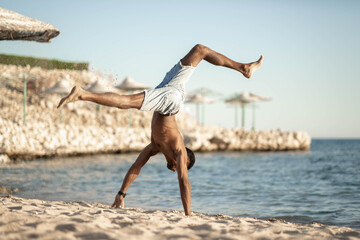 Fototapeta na wymiar Handsome black young man topless dancing break dance, jumping, handstand, spinning, posing upside down on sand beach 
