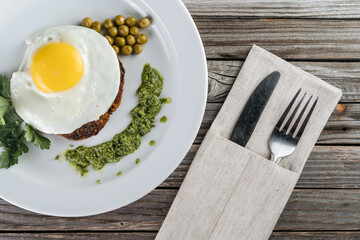Scrambled eggs with meat on a plate, garnished with green peas and pesto sauce. Nearby are a knife and a fork.