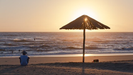 Beach Shoreline Man Sitting By Umbrella Silhouetted Watching Sunrise Over Ocean Sea Horizon.