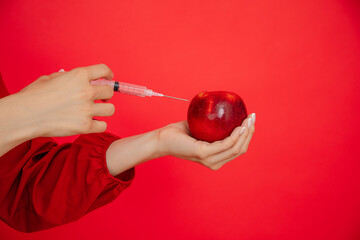 Cropped woman hand with syringe apply injection to fresh juicy red apple in red studio. Botox,...