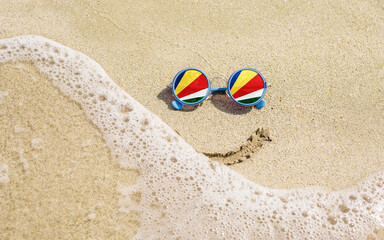 Fototapeta na wymiar Sunglasses with flag of Seychelles on a sandy beach. Nearby is a sea lightning and a painted smile. The concept of a successful vacation in the resorts of the Seychelles.